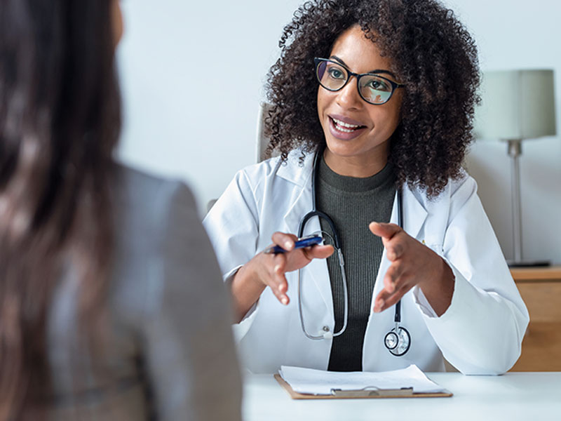Una doctora con cabello rizado y gafas, con un estetoscopio alrededor del cuello, está sentada en un escritorio, entablando una conversación con una paciente mientras sostiene un bolígrafo y gesticula con las manos.